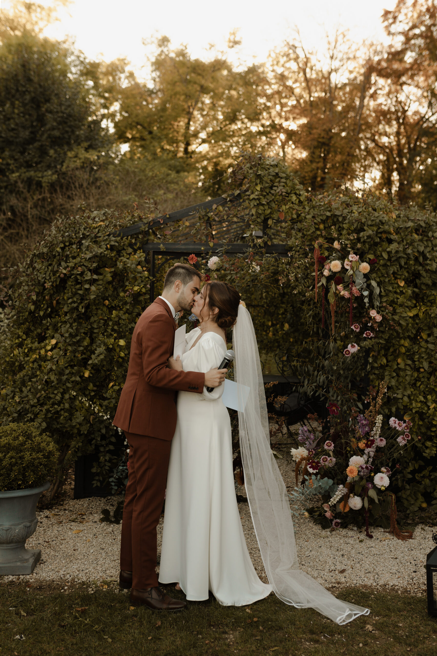 Photographie de mariage en couleur au Domaine de La Closeraie, près de Reims. Un couple s'embrasse passionnément pendant sa cérémonie laïque en extérieur, devant un kiosque contemporain noir, dans un parc verdoyant en Champagne.