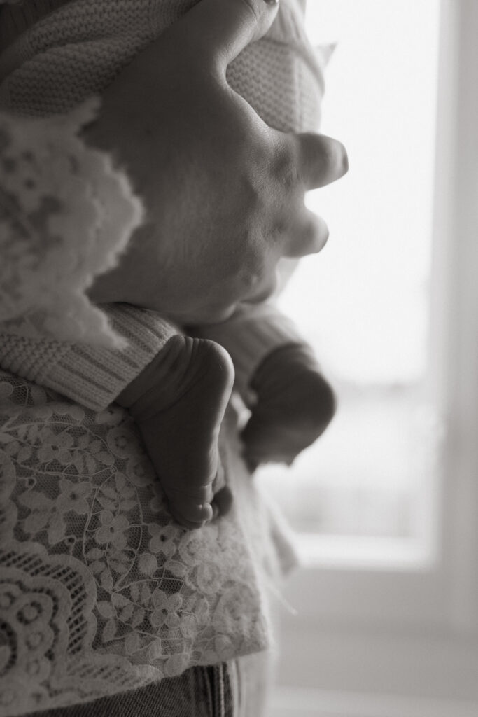 Photo de naissance en noir et blanc à Reims : gros plan d'une main tenant les fesses d'un bébé, petits pieds pendants. Lumière naturelle en contre-jour.