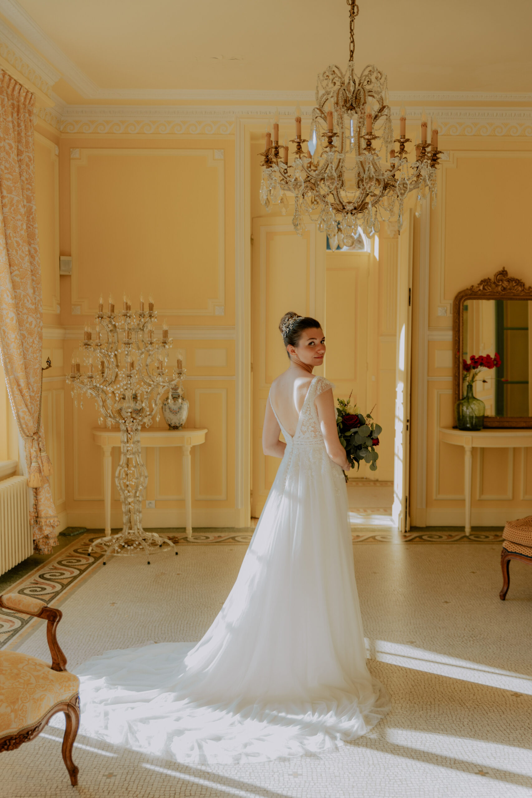 Portrait en pied de la mariée dans le salon jaune du Château de Droupt-Saint-Basle, lustre luxe et rayons de soleil.