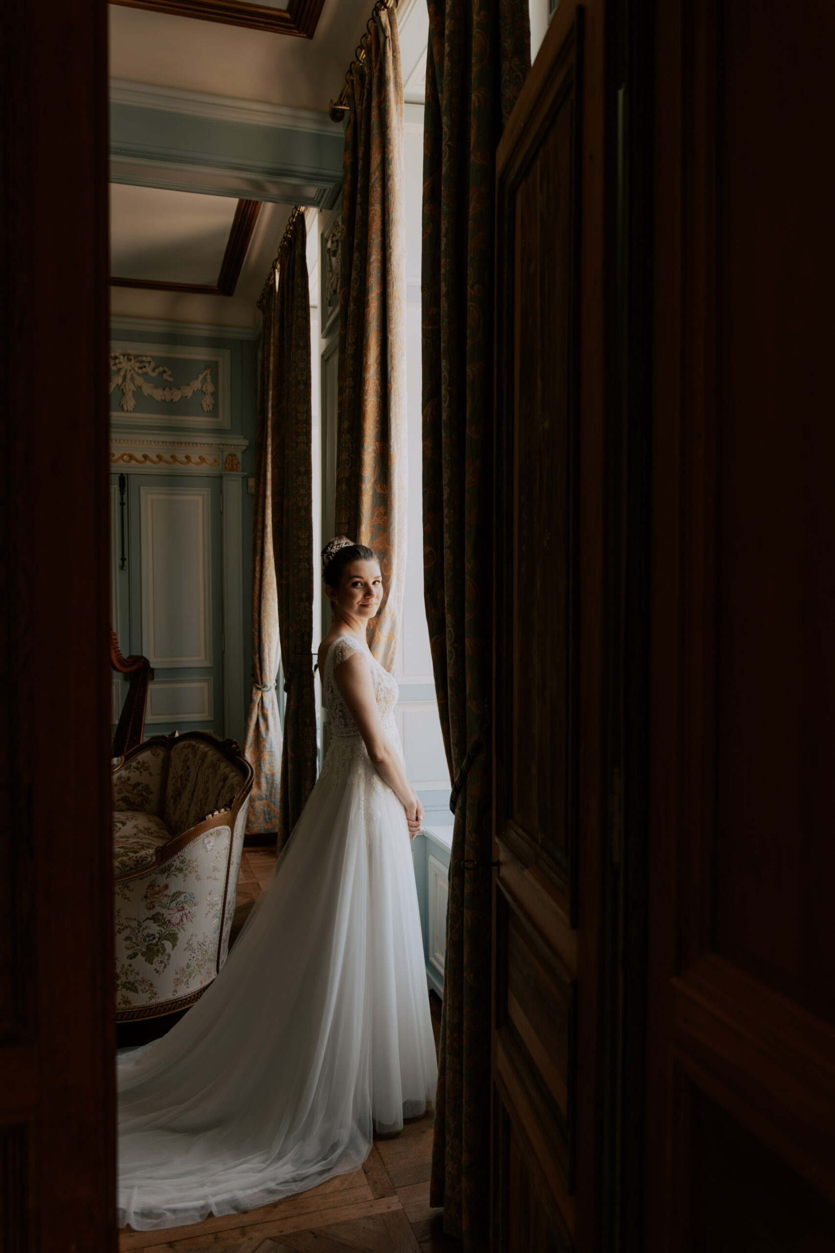 Portrait en pied de la mariée face à la fenêtre du salon bleu, jeu de lumière clair-obscur.