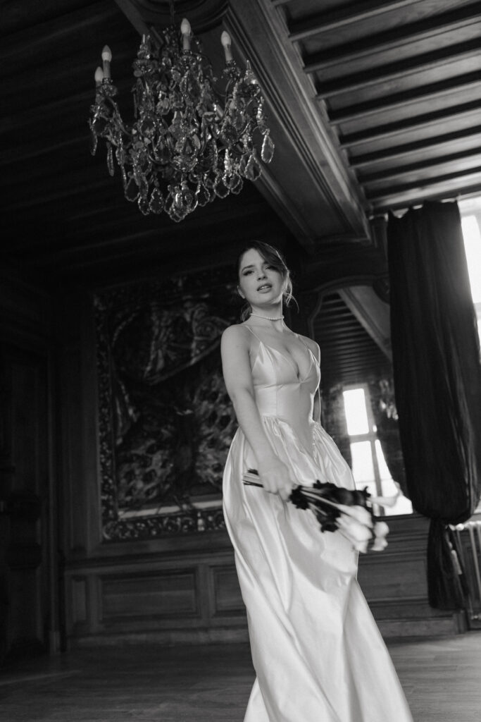 Photo de mariage éditorial en noir et blanc d'une mariée dansant et regardant l'objectif, au Château de Vitry-la-Ville dans la Marne. Effet de mouvement et voile volant. Photographe mariage Champagne-Ardenne.