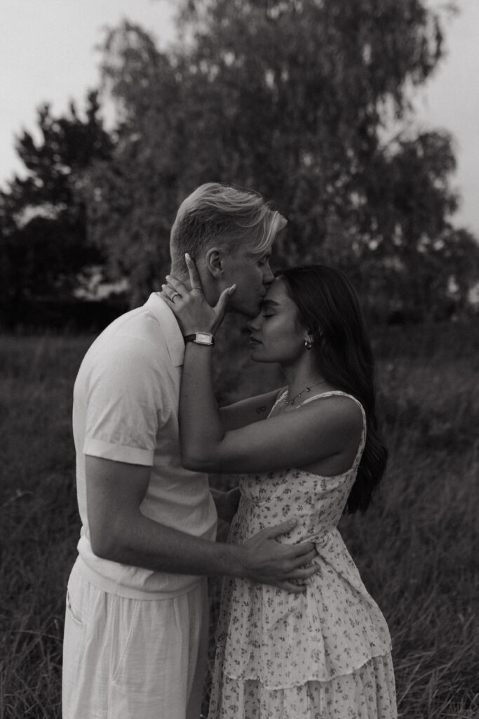 Portrait intime en noir et blanc d'un couple s'embrassant sur le front. La femme ferme les yeux et tient la nuque de son compagnon, lors d'une séance photo près de Reims, Champagne-Ardenne.