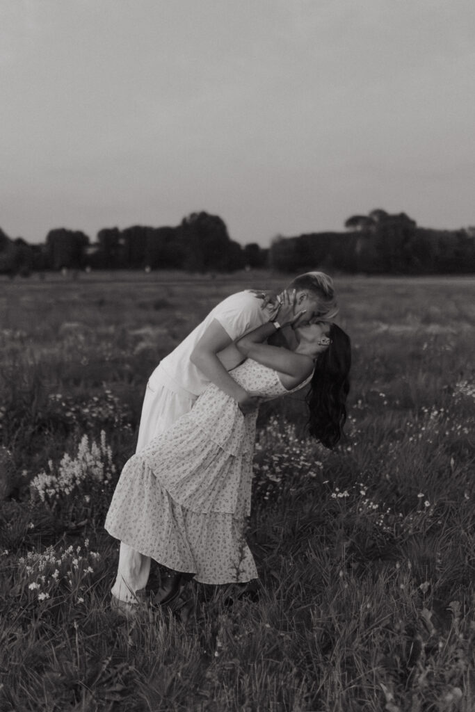 Photographie en noir et blanc en pied (plan large) d'un couple lors d'une séance photo à la Blue Hour près de Reims, Champagne-Ardenne. L'homme soutient sa femme dans une pose balancée pour un baiser passionné (deep kiss) au milieu d'un champ de fleurs sauvages.
