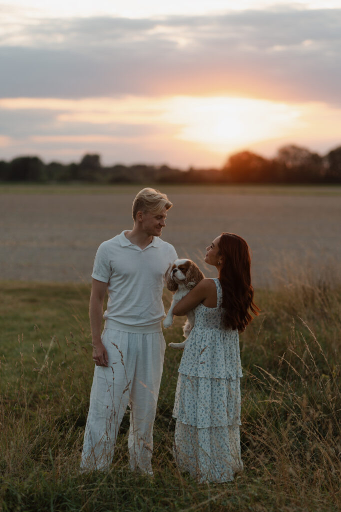 Photographie de couple au coucher du soleil à Prunay lors d'une séance engagement près de Reims. Ils se regardent amoureusement, entourés de hautes herbes dorées, tandis que leur petit chien Cavalier King Charles est à leurs côtés.