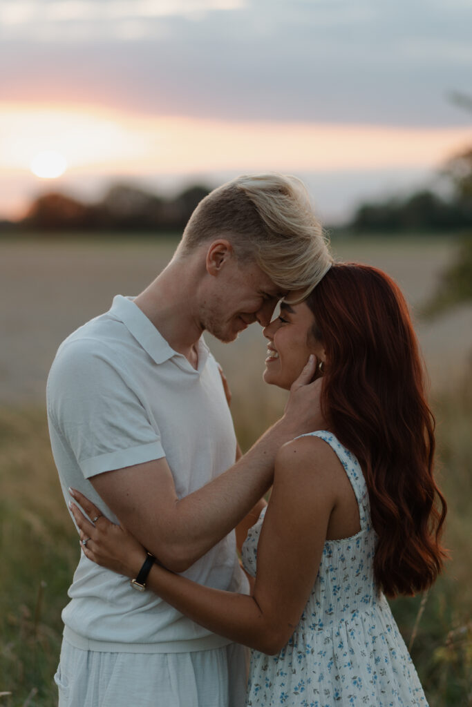 Portrait rapproché d'un couple riant aux éclats lors d'une séance engagement à Prunay, près de Reims. Ils sont face à face dans un champ de hautes herbes dorées au coucher du soleil.