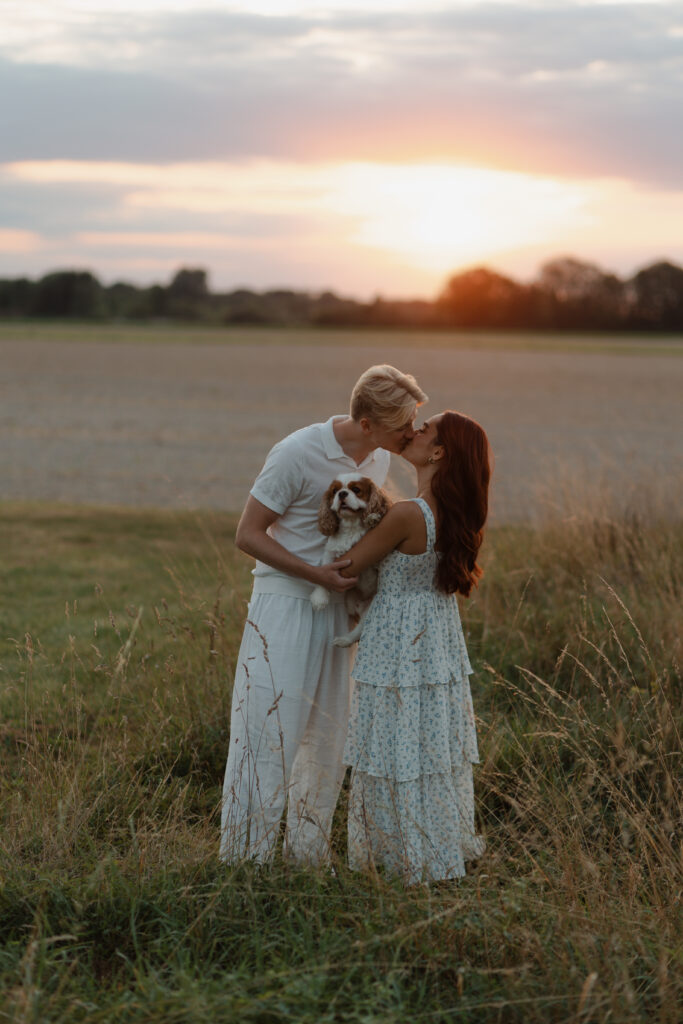 Couple s'embrassant dans un champs au coucher du soleil avec leur cavalier king charles dans les bras, lors d'une séance couple à Prunay près de Reims.
