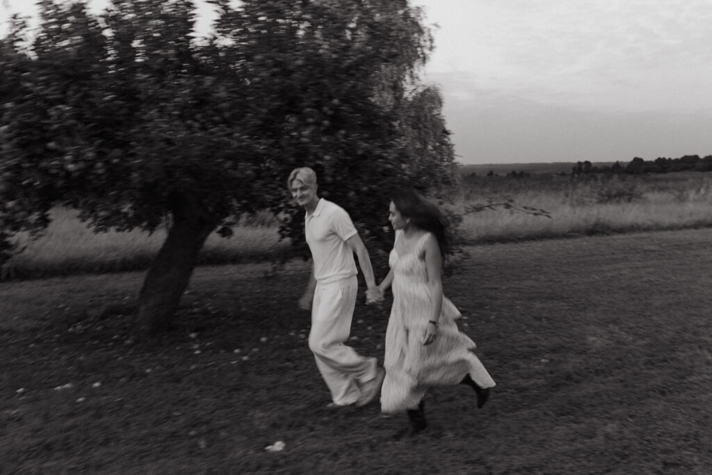 Photographie artistique en noir et blanc avec un fort flou de mouvement d'un couple courant et se regardant. Séance engagement à Prunay (Reims) au pied d'un pommier, capturant l'énergie et la vitesse.