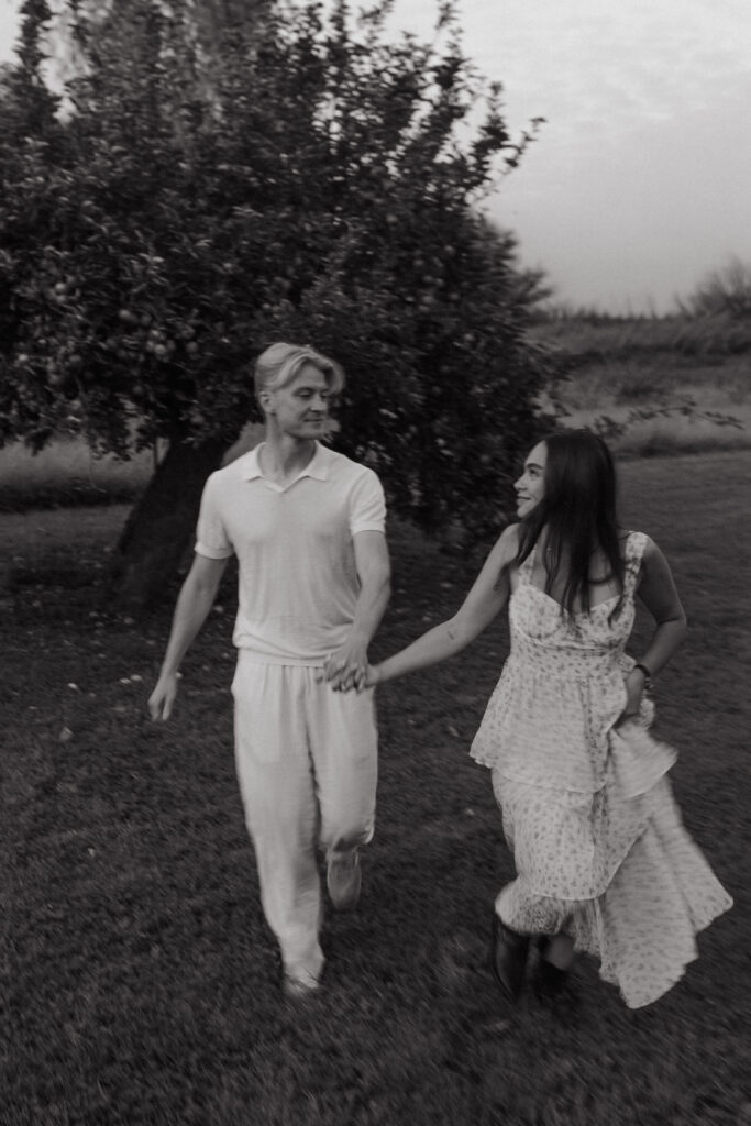 Photographie artistique en noir et blanc avec flou de mouvement lors d'une séance engagement à Prunay, près de Reims. Un couple court vers l'appareil photo en se regardant, créant un effet de vitesse dynamique au pied d'un pommier.