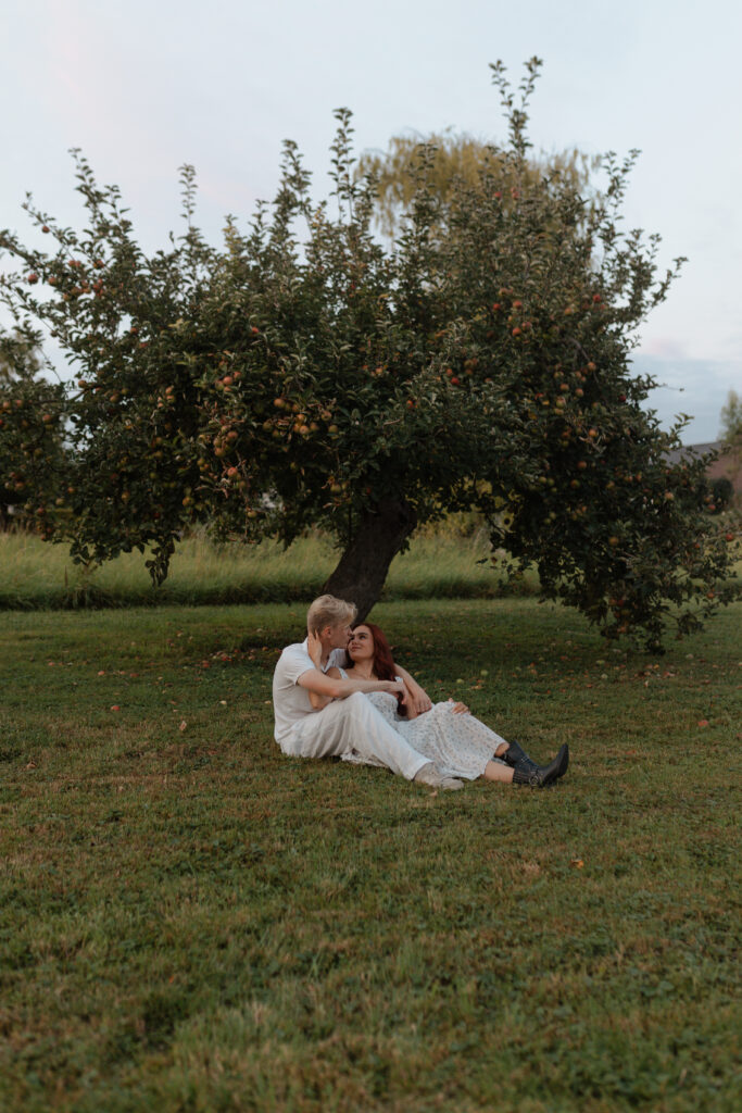 Photographie verticale en plan large d'un couple assis au pied d'un pommier à Prunay. Le cadrage met en valeur l'immensité du paysage champenois et la nature environnante lors d'une séance engagement près de Reims.