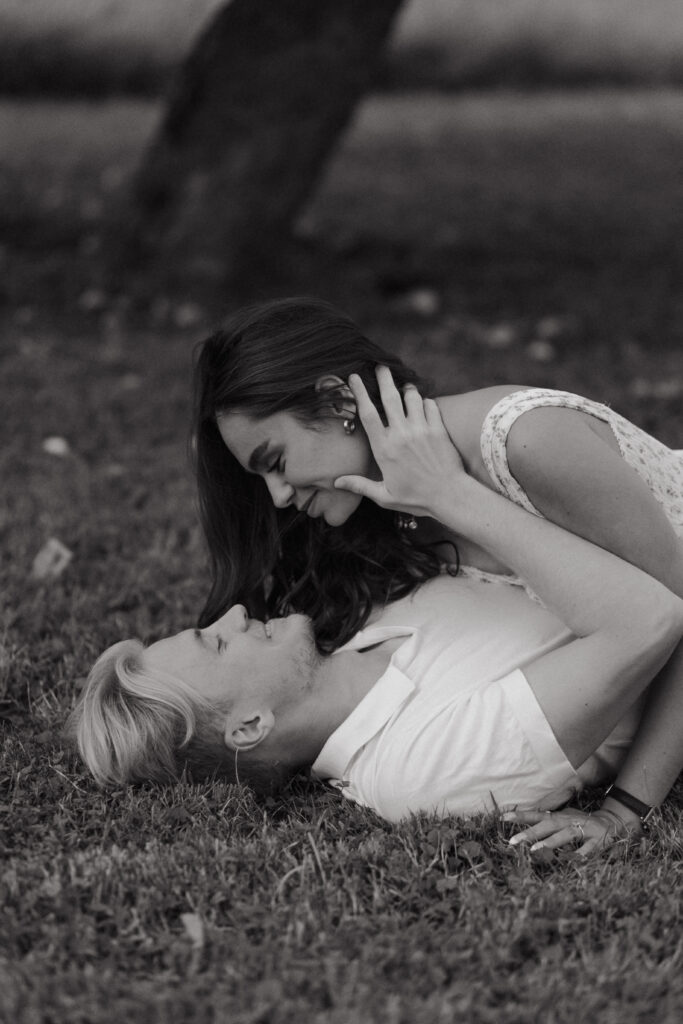 Portrait rapproché artistique en noir et blanc d'un couple allongé dans l'herbe au pied d'un pommier à Prunay. La femme est au-dessus de son fiancé et ils se regardent avec intensité lors d'une séance engagement près de Reims.