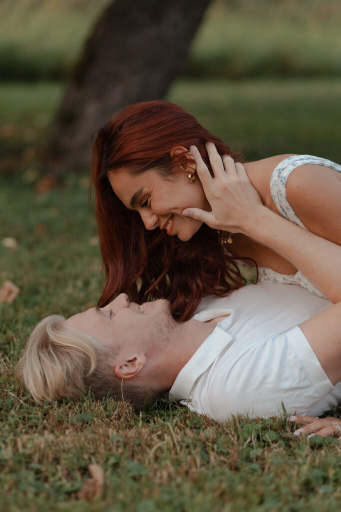 Portrait rapproché d'un couple allongé dans l'herbe au pied d'un pommier à Prunay. La femme est au-dessus de son fiancé et ils se regardent avec complicité lors d'une séance engagement près de Reims.