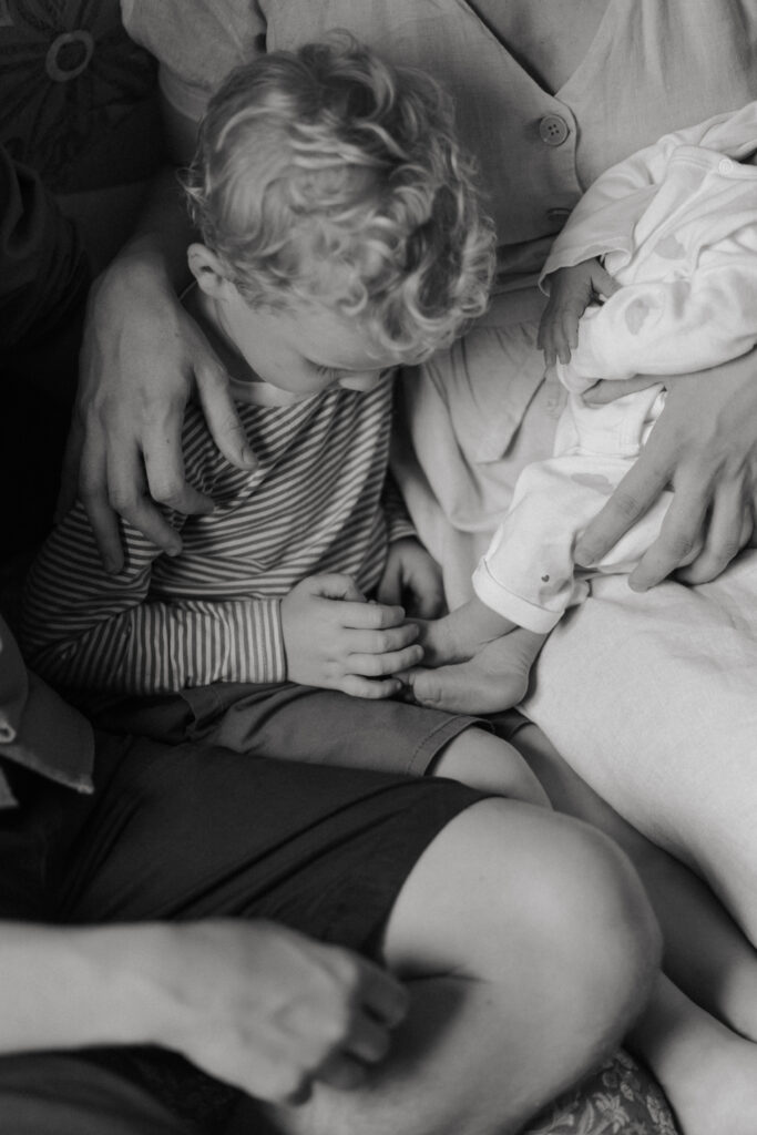 Photo en noir et blanc d'un bébé tenu dans les bras de sa mère, grand frère caressant les pieds du nouveau-né à côté. Portrait de famille chaleureux dans la Marne.