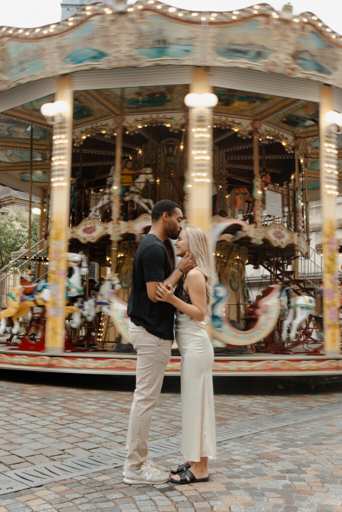 Séance couple en couleurs dans l'hypercentre de Reims. Un homme embrasse sa femme sur le front devant le carrousel en mouvement de flou. Capturé par votre photographe à Reims.