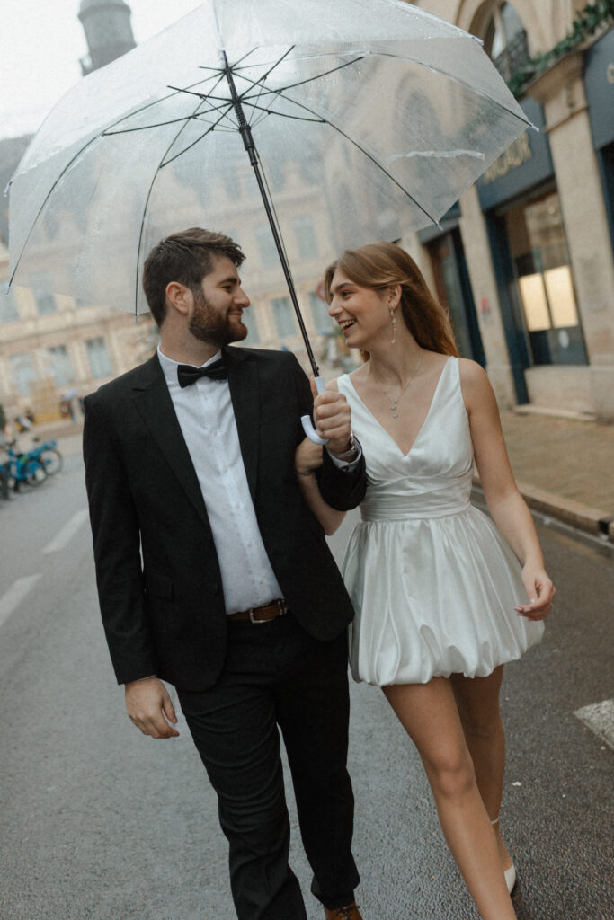Couple de mariés complices marchant sous la pluie sous un parapluie transparent en centre-ville de Reims.