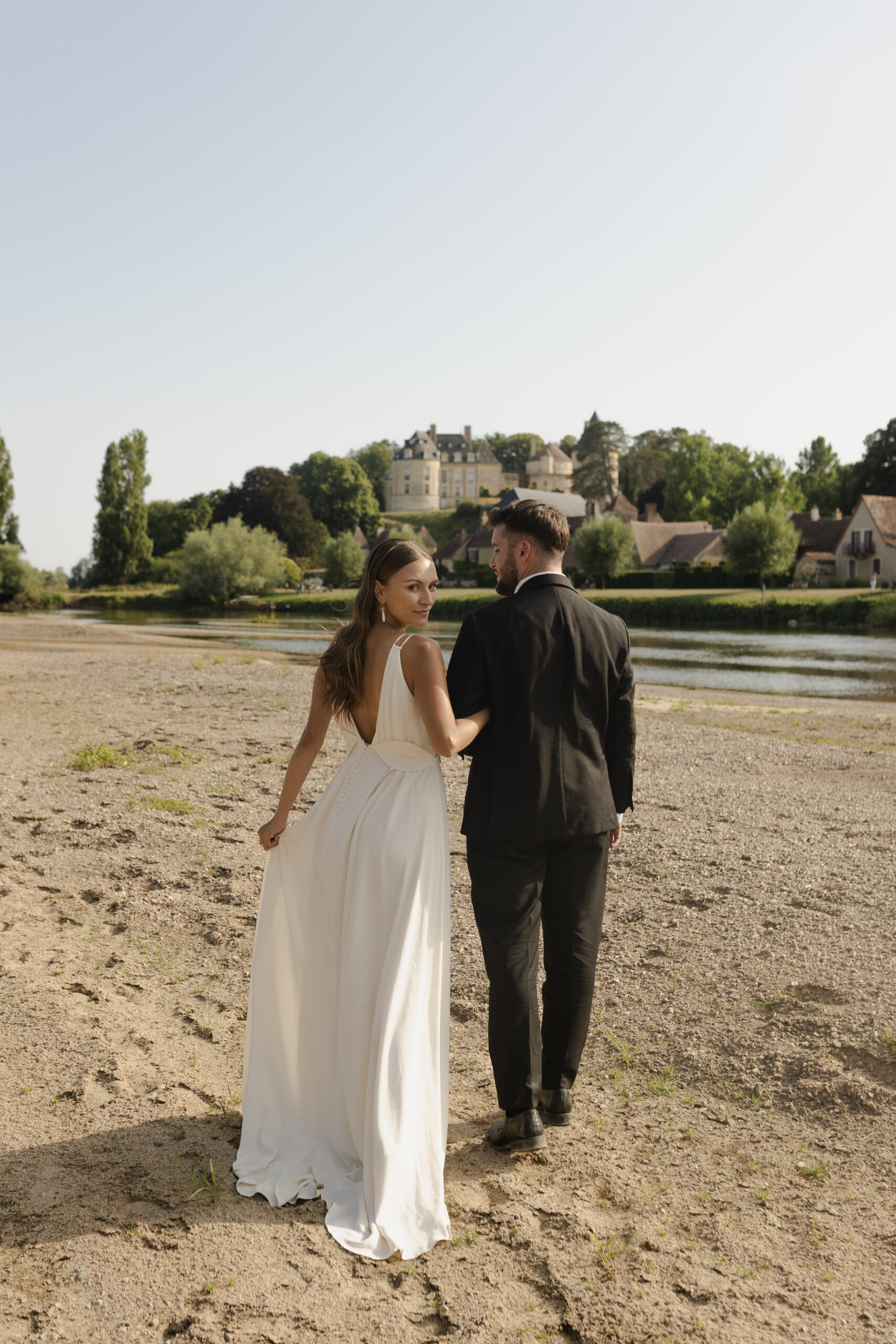 Mariés marchant sur une plage de sable avec vue sur un château en Dordogne.