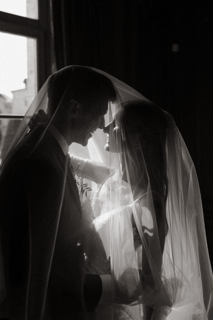Silhouette de couple de mariés sous un voile en contre-jour près d'une fenêtre.