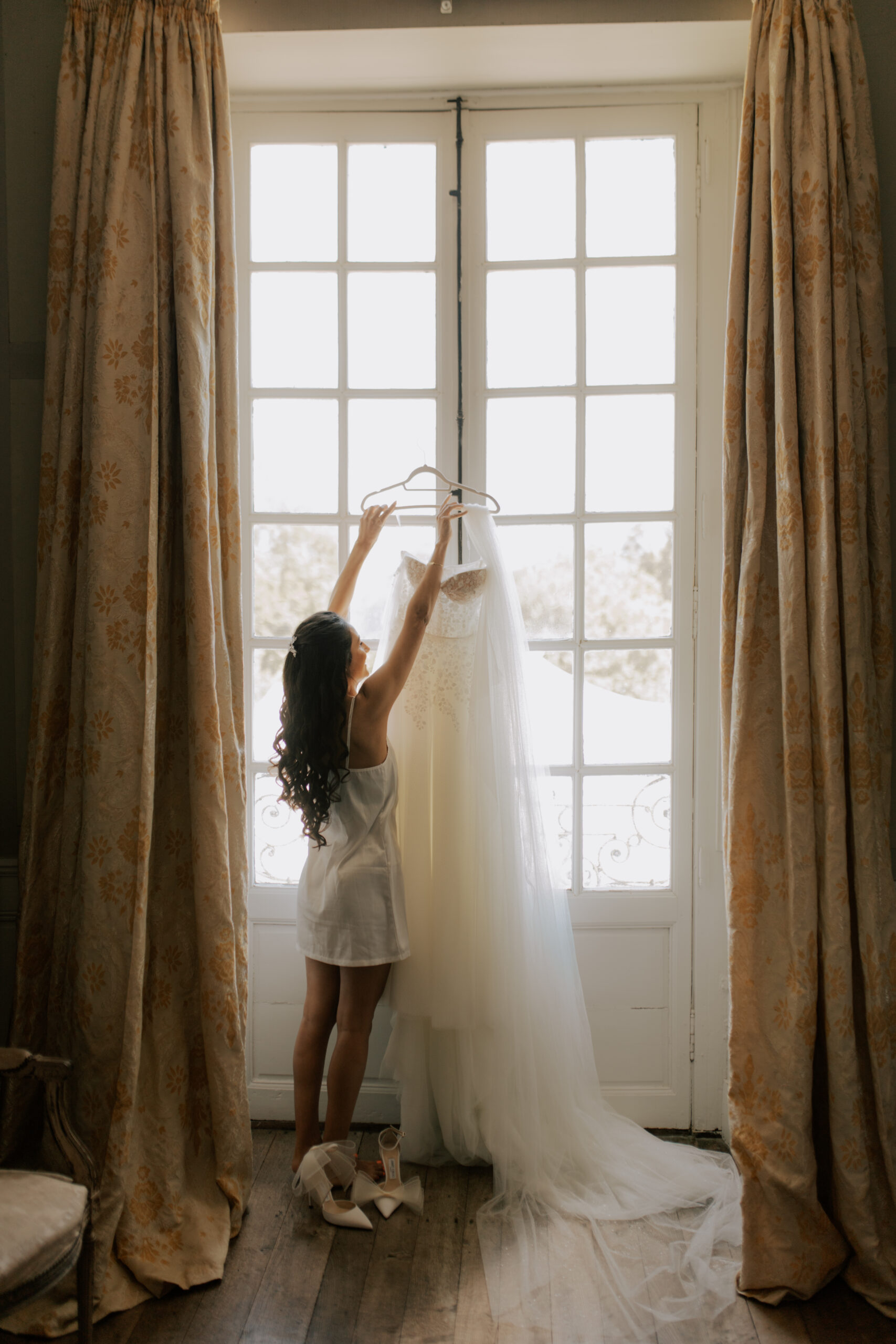 Robe de mariée suspendue sur un cintre devant une fenêtre du Château de Mairy à contre-jour.