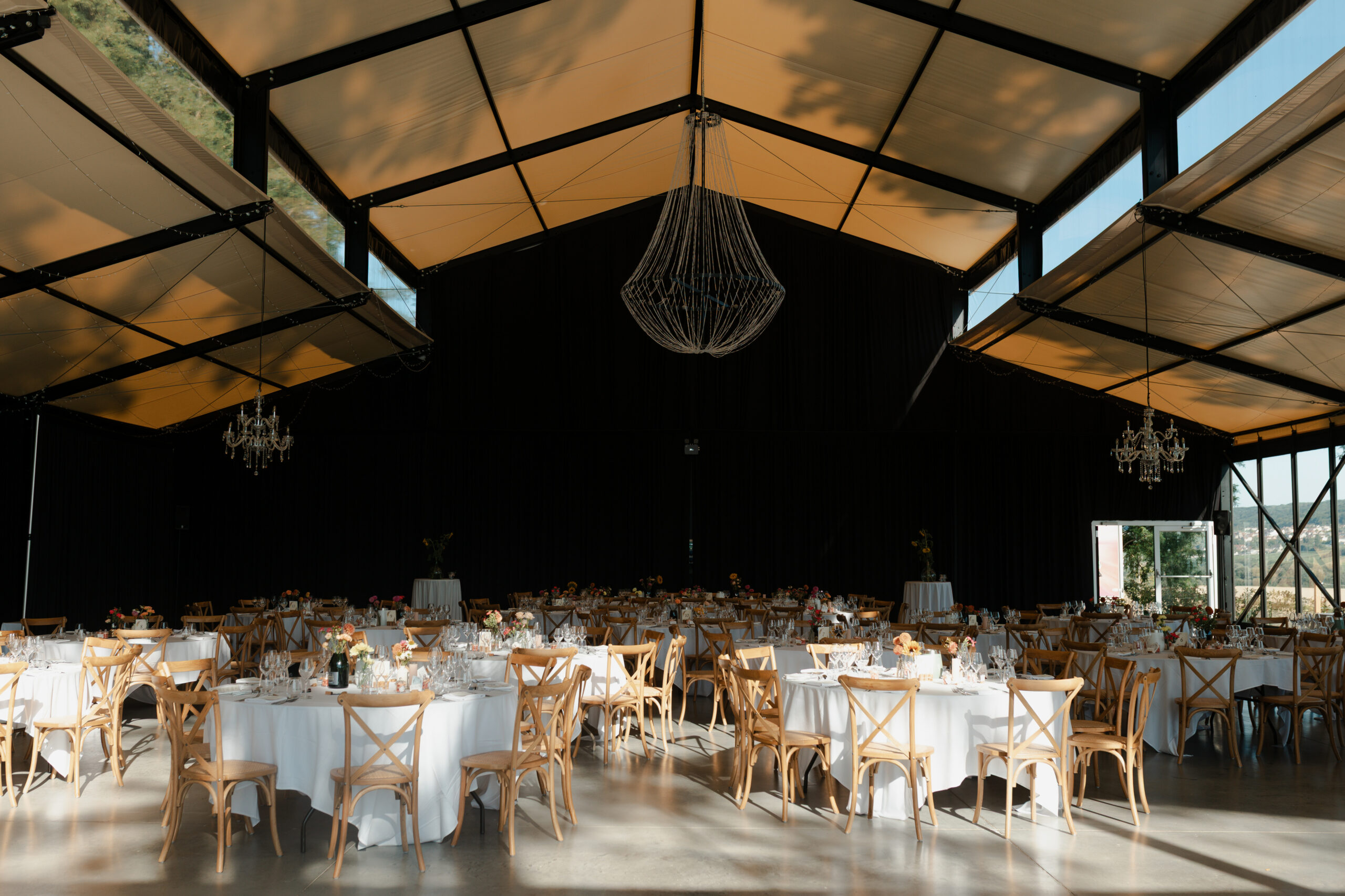 Vue d'ensemble de l'orangerie du Prieuré de Binson décorée pour un mariage. Plafond haut et lustre élégant visibles.