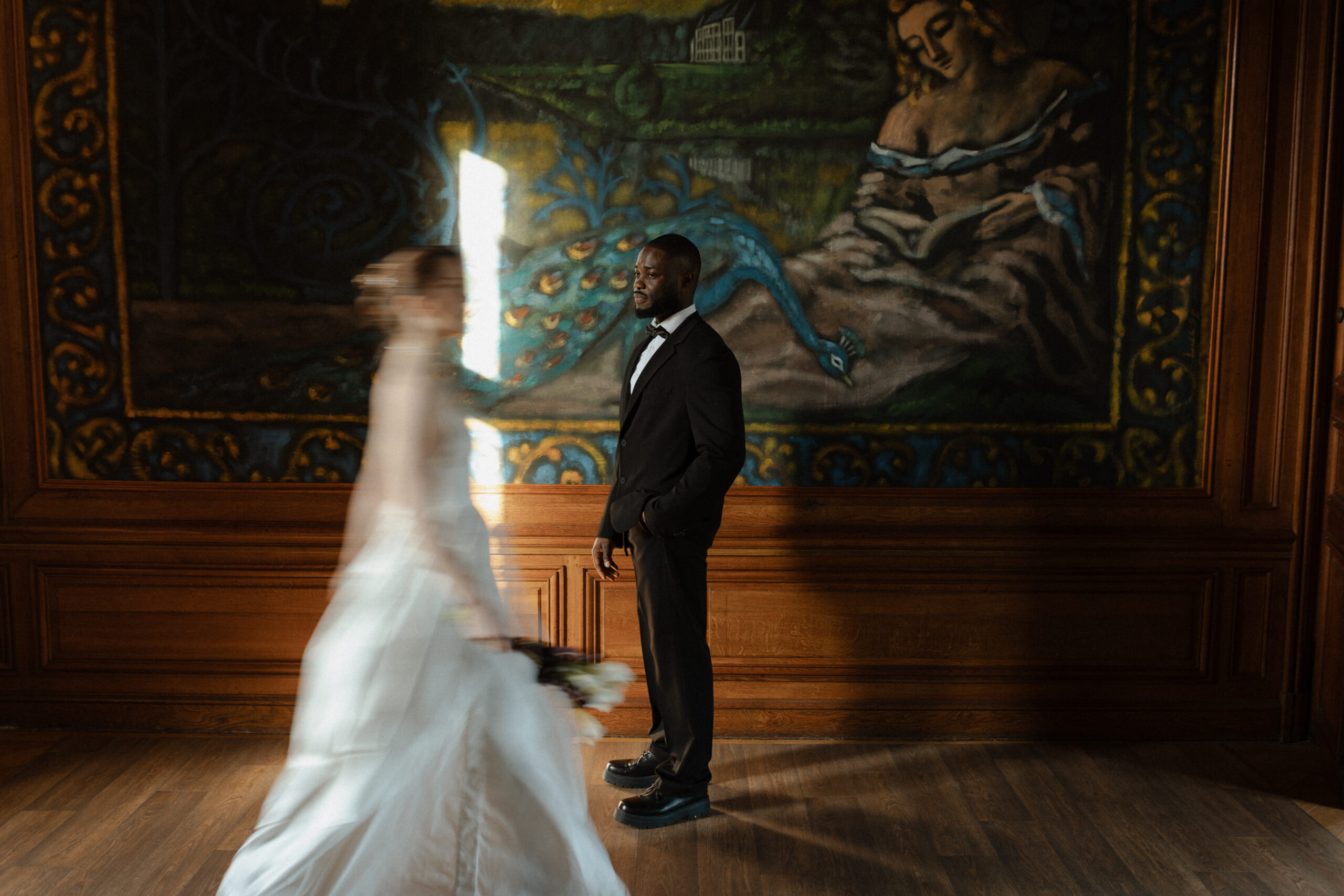Photo artistique du marié devant une tapisserie d'époque, la mariée passe au premier plan avec un effet de flou.