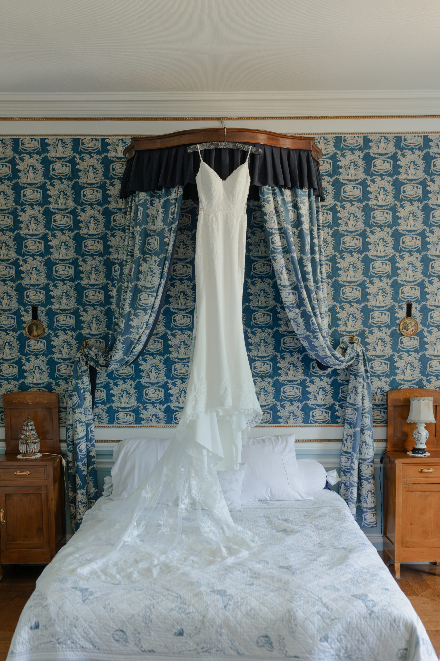 Robe de mariée suspendue au-dessus du lit dans une chambre d'époque avec tapisserie bleue et blanche.
