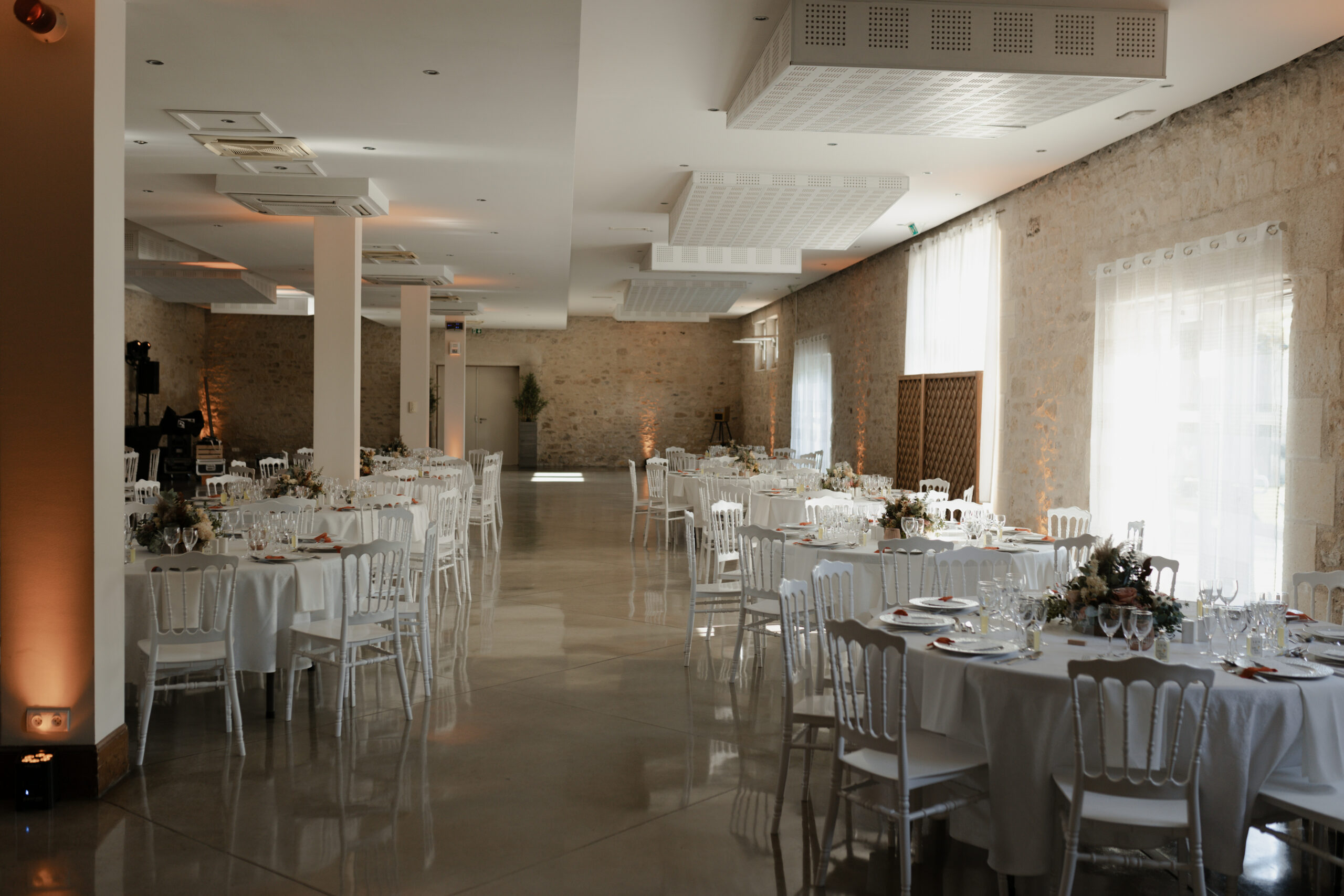 Salle intérieure du domaine Les Pommerieux avec murs en pierre apparente et tables de mariage dressées.