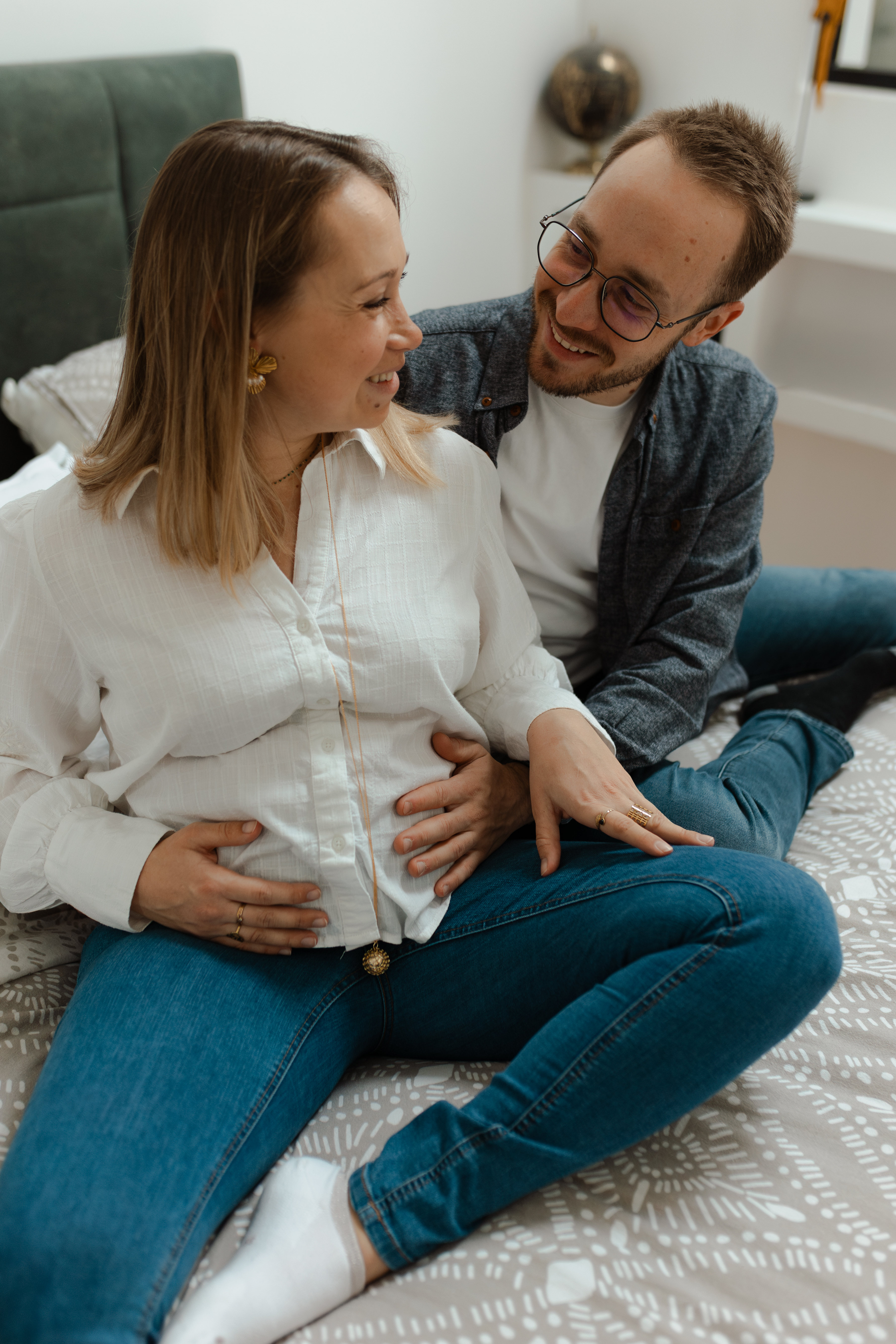 Couple assis sur leur lit se regardant avec complicité, mains posées sur le ventre de grossesse de la maman. Séance photo grossesse lumineuse à Reims.
