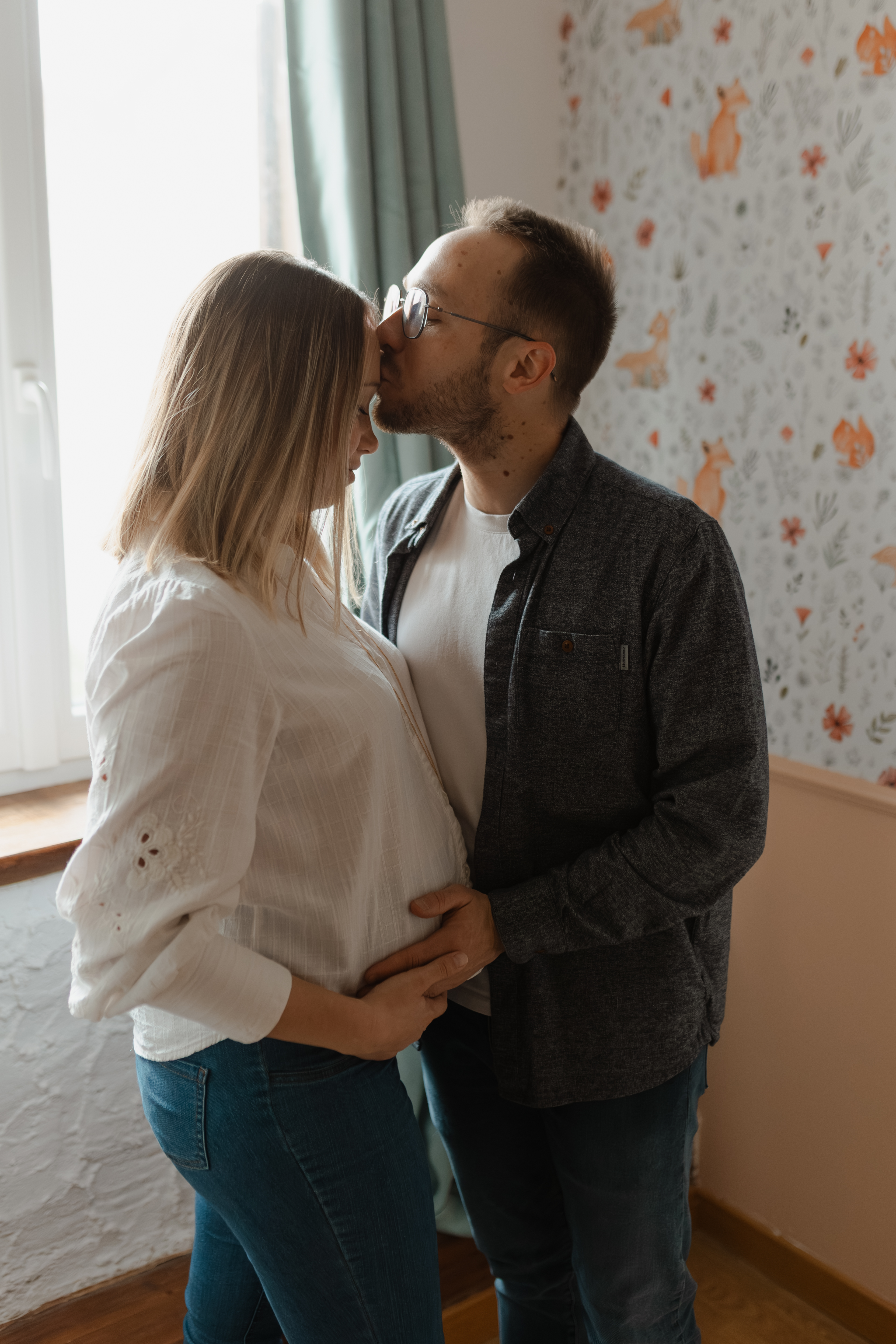 Couple en contre-jour devant une fenêtre : le futur papa embrasse le front de la maman, mains sur le ventre de grossesse. Séance photo à Reims.