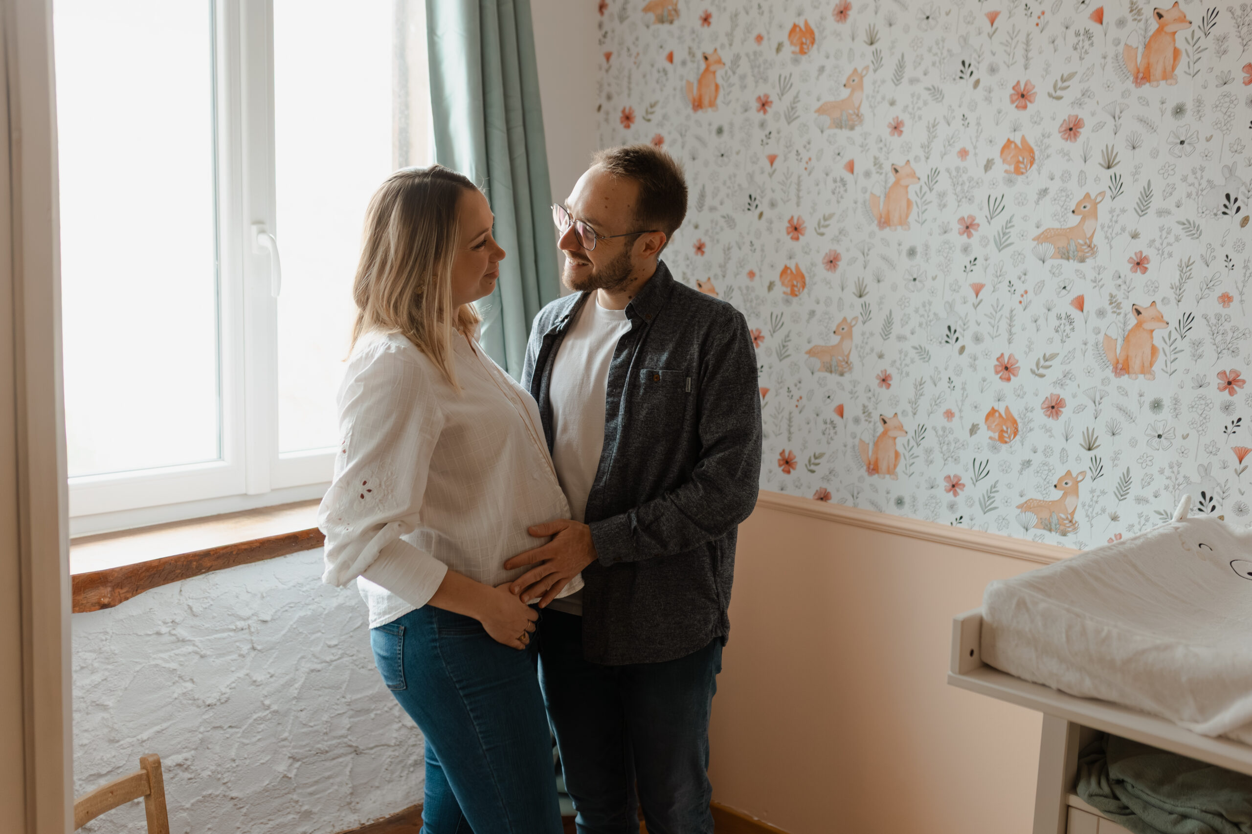 Séance photo grossesse dans la chambre de bébé près de Reims : couple complice se regardant tendrement, mains posées sur le ventre arrondi.