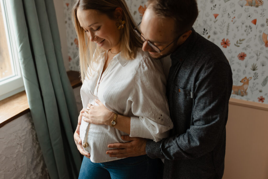Couple dans la chambre de bébé à Reims : ils regardent tendrement le ventre de grossesse. Séance grossesse douce.