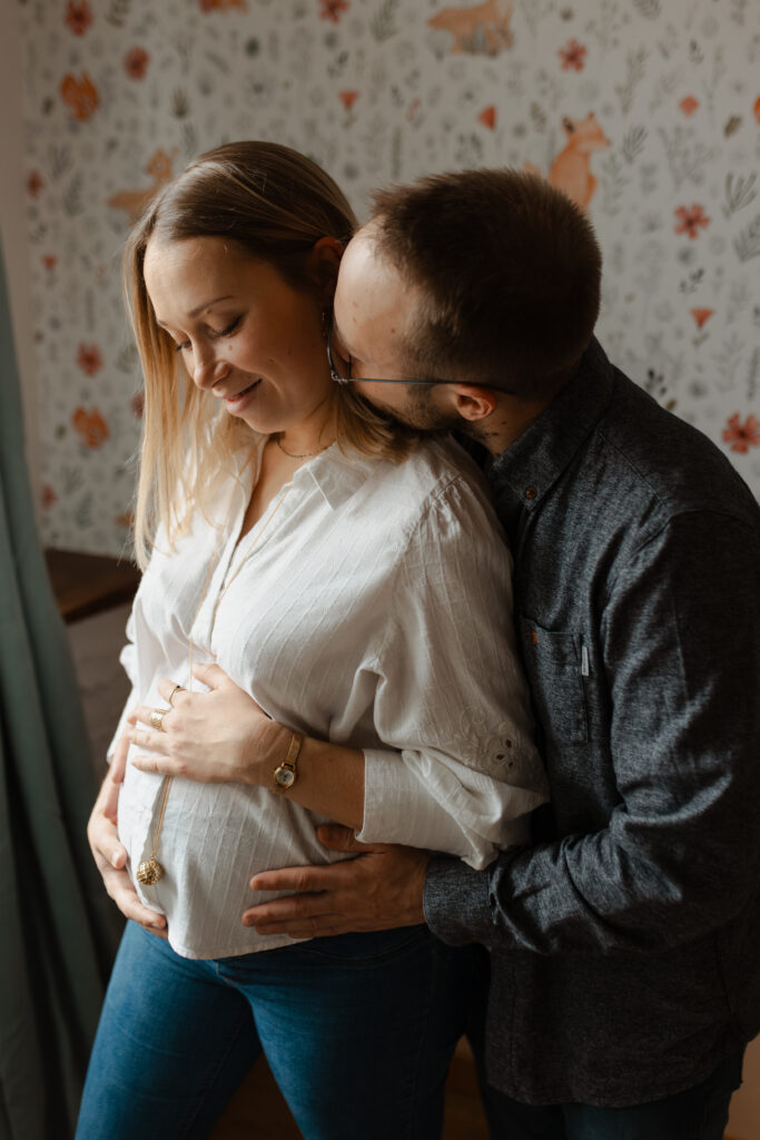 Futur papa embrassant le cou de la future maman dans la chambre de bébé à Reims. Photo grossesse intime.