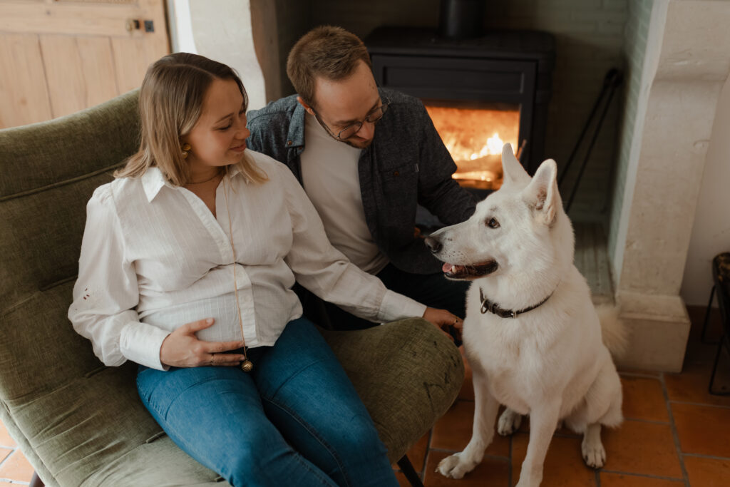 Couple de futurs parents avec leur chien blanc sur un fauteuil devant la cheminée à Reims.
