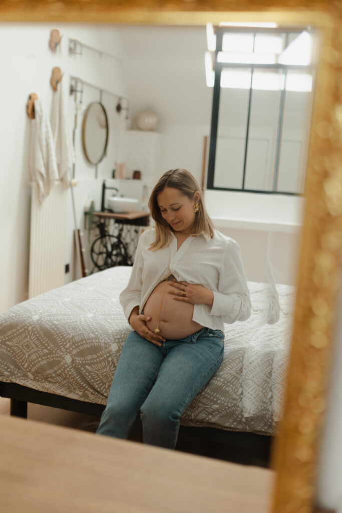Portrait d'une future maman assise sur son lit, vue en reflet dans le miroir. Elle regarde tendrement son ventre de grossesse. Photographe à Reims.
