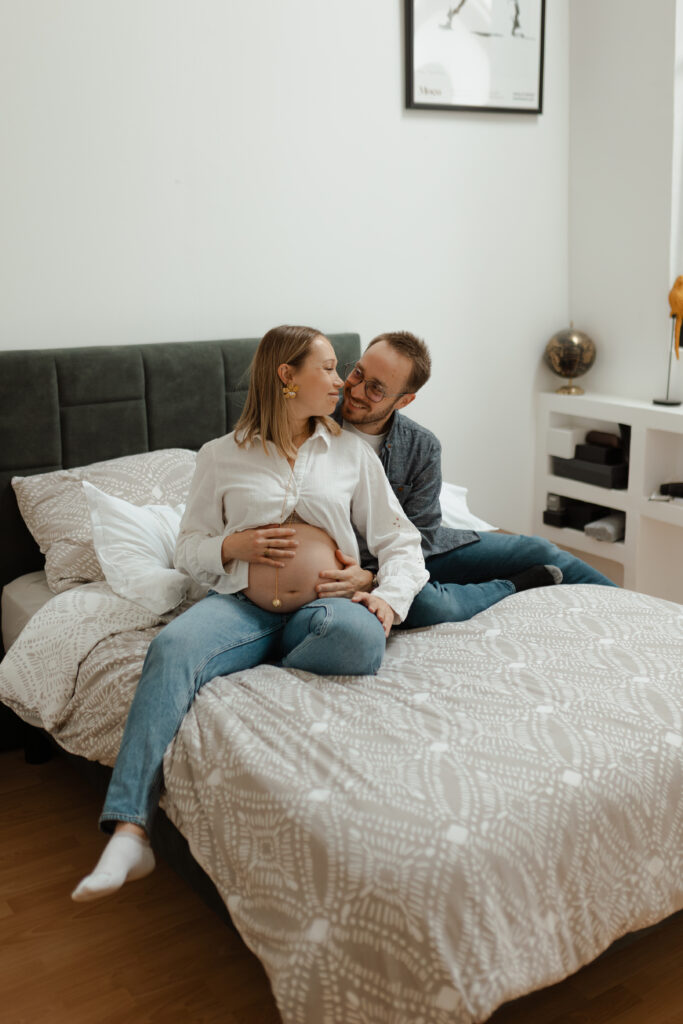 Séance photo grossesse en plan large à Reims : couple complice assis sur leur lit, mains sur le ventre, dans une chambre lumineuse et douce.