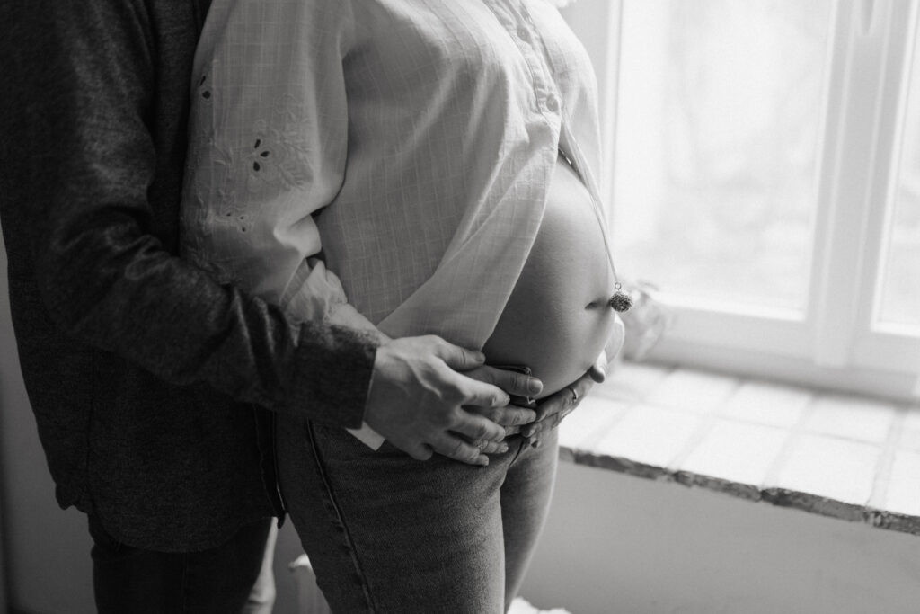 Photo en noir et blanc : mains des parents posées délicatement sous le ventre de grossesse.