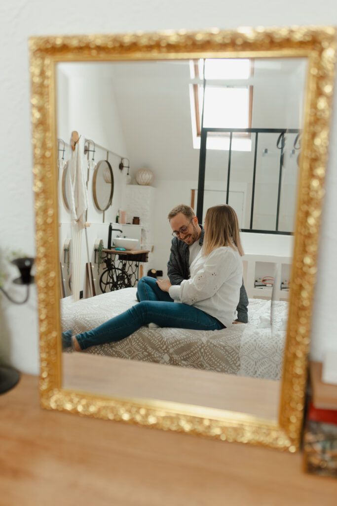 Séance photo grossesse à domicile près de Reims : reflet dans le miroir du visage souriant du futur papa, vue sur la chambre.