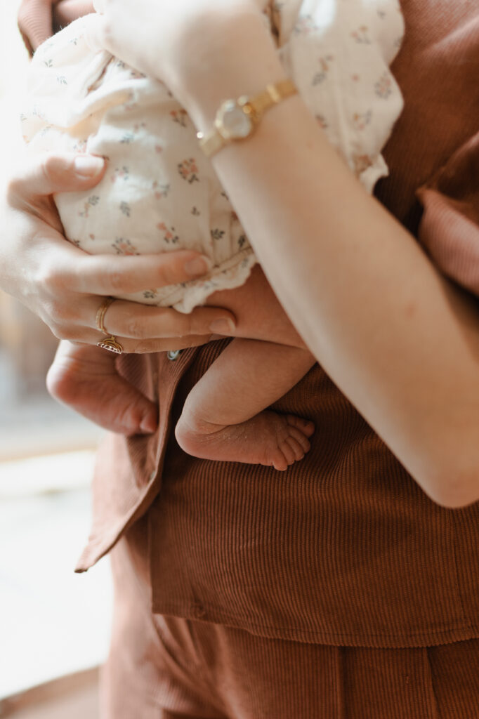 Photo de naissance à Reims : gros plan des petits pieds d'un bébé porté par sa mère qui le soutient par les fesses. Style doux et lumineux.