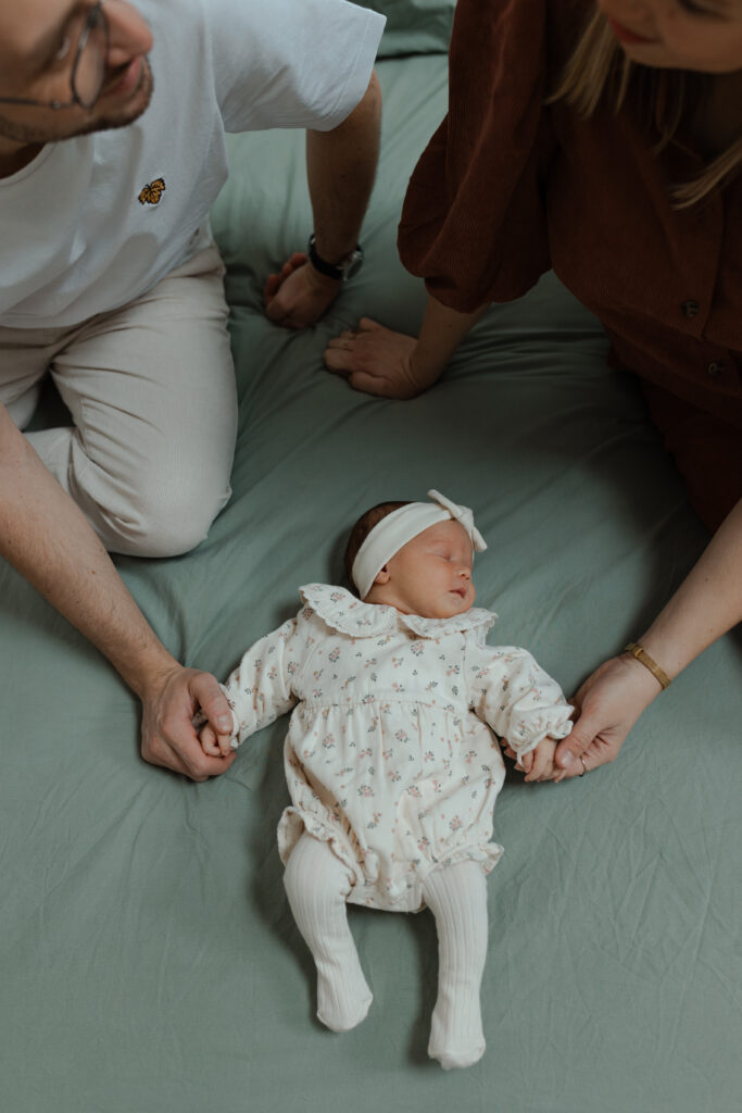 Parents tenant chacun une main de leur bébé allongé sur le lit.