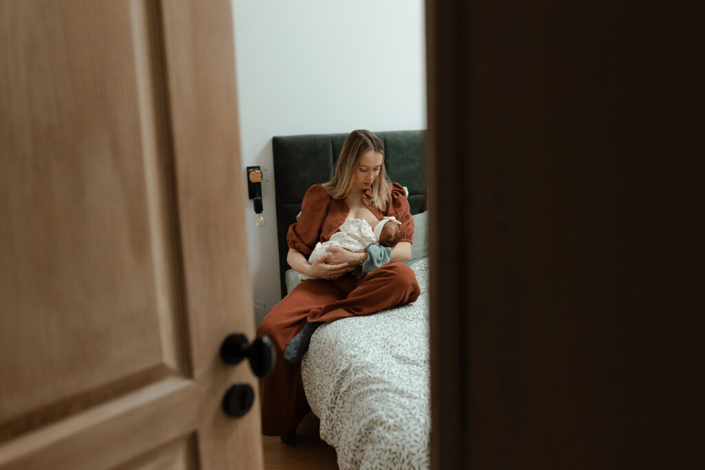 Maman allaitant son nouveau-né assise sur son lit, vue par l'entrebâillement d'une porte à Reims.