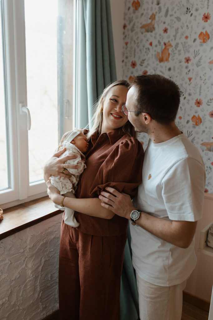 Couple complice dans la chambre de bébé, papa enlaçant la maman qui porte le bébé.