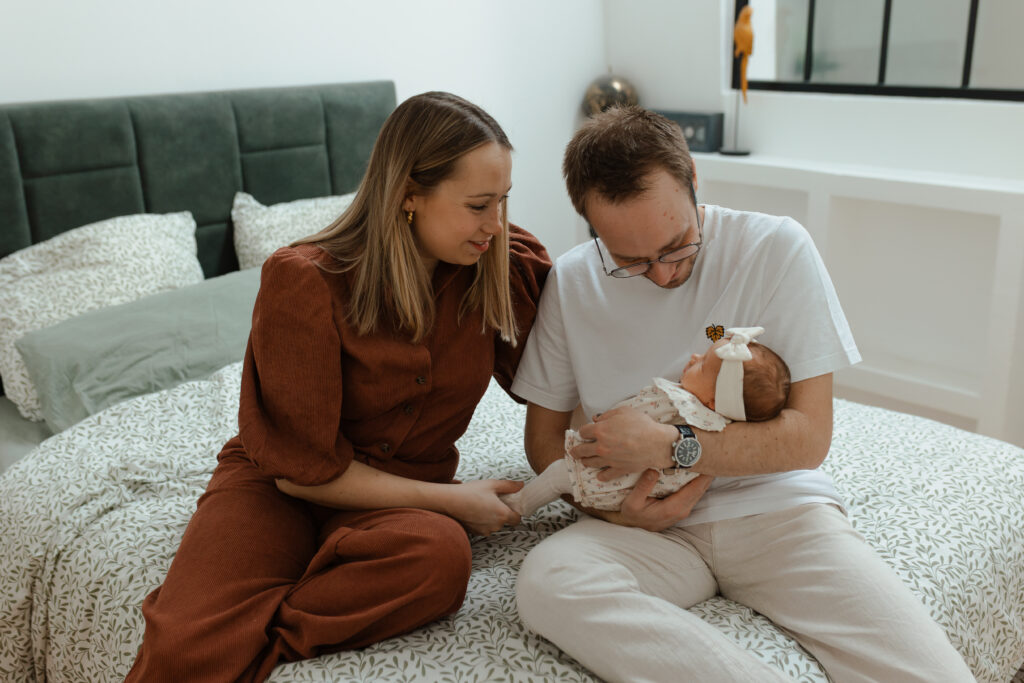 Couple de parents souriants regardant leur nouveau-né dans les bras du papa, assis sur leur lit à Reims.